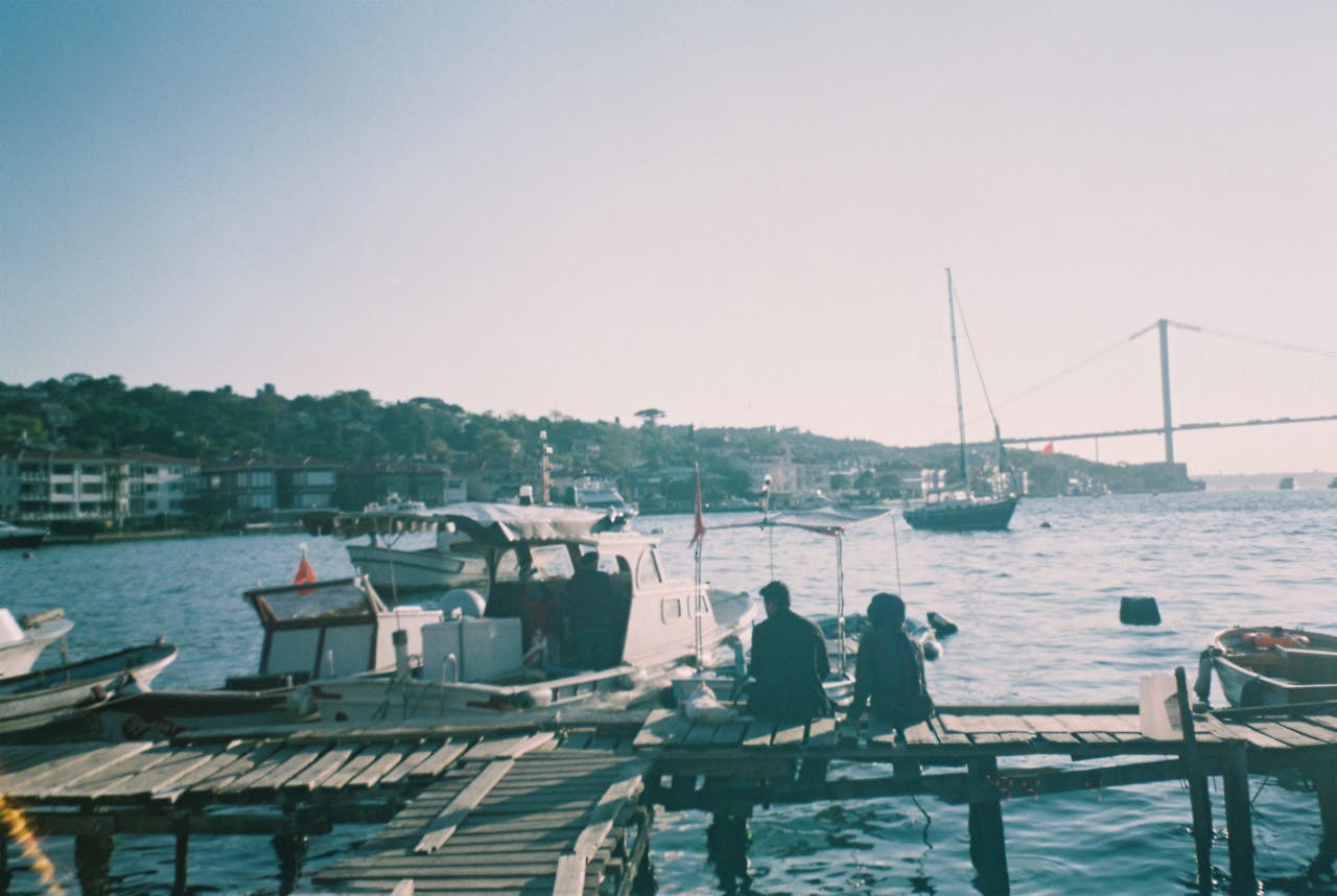 A peaceful harbor scene with boats and a bridge at sunset, two people on the dock.